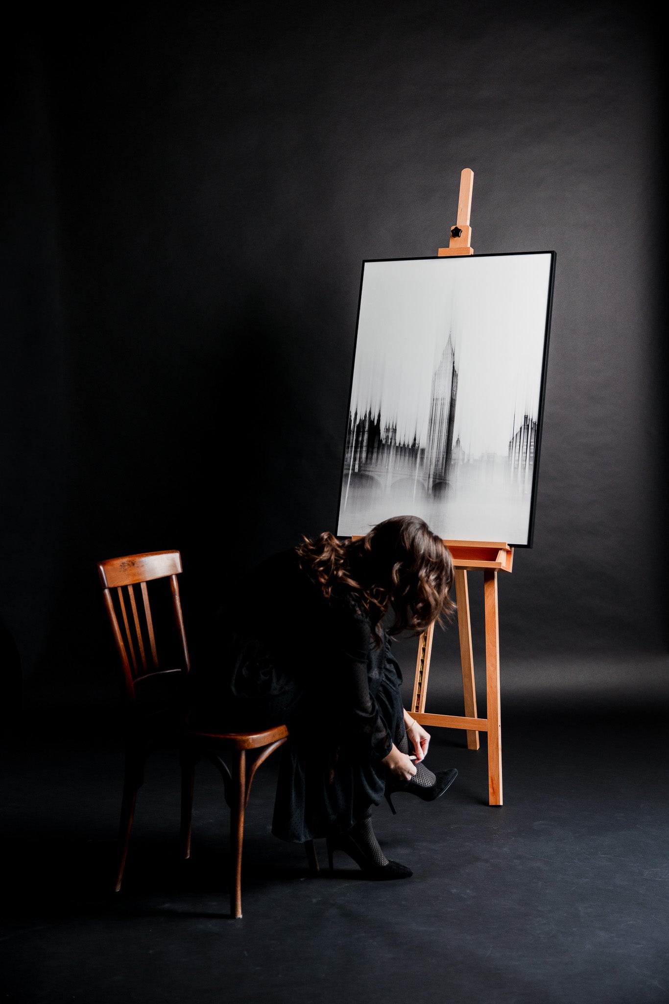 Schwarz-weiss Fine Art Fotografie „Ben Whispered“ von Motel June auf einer Staffelei, kunstvolle Aufnahme des Big Ben Towers in London, im Studio sitzt eine junge Frau auf einem braunen Stuhl und schließt ihre eleganten High Heels vor schwarzem Hintergrund.