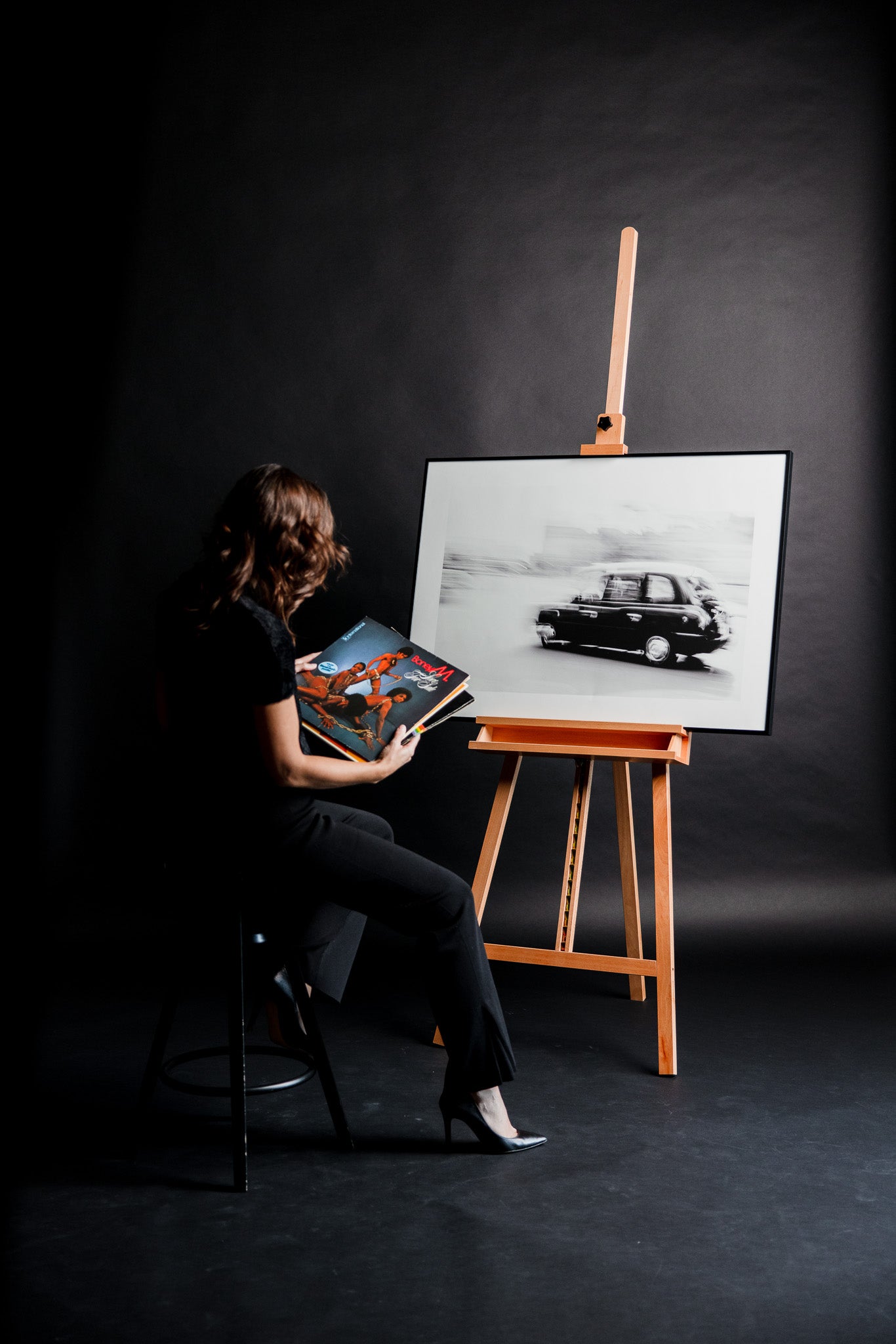 Schwarz-weiss Fotokunst „Churchill“ von Motel June auf einer Staffelei vor schwarzem Studiohintergrund, London Panning Shot mit englischem Taxi, links ein alter brauner Stuhl, darauf sitzt eine elegante junge Frau in schwarzen Kleidern und wählt Vinyl-Schallplatten aus.