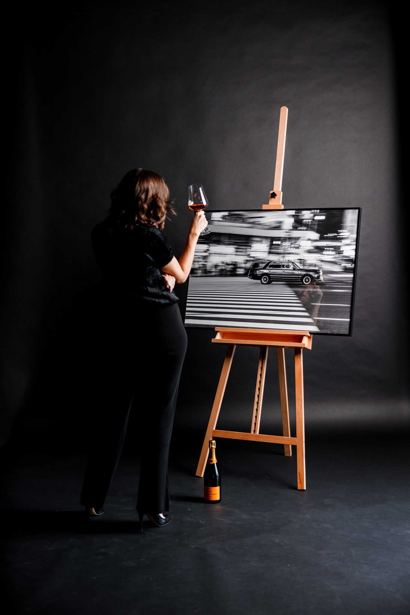 Schwarz-weiss Kunstfotografie „Guilty“ von Motel June auf einer Staffelei, japanisches Taxi in Tokyo bei einem dynamischen Panning Shot mit verzerrten Zebrastreifen, elegante Frau in schwarzen Kleidern steht davor und genießt das Bild mit einem Glas Champagner in der Hand.