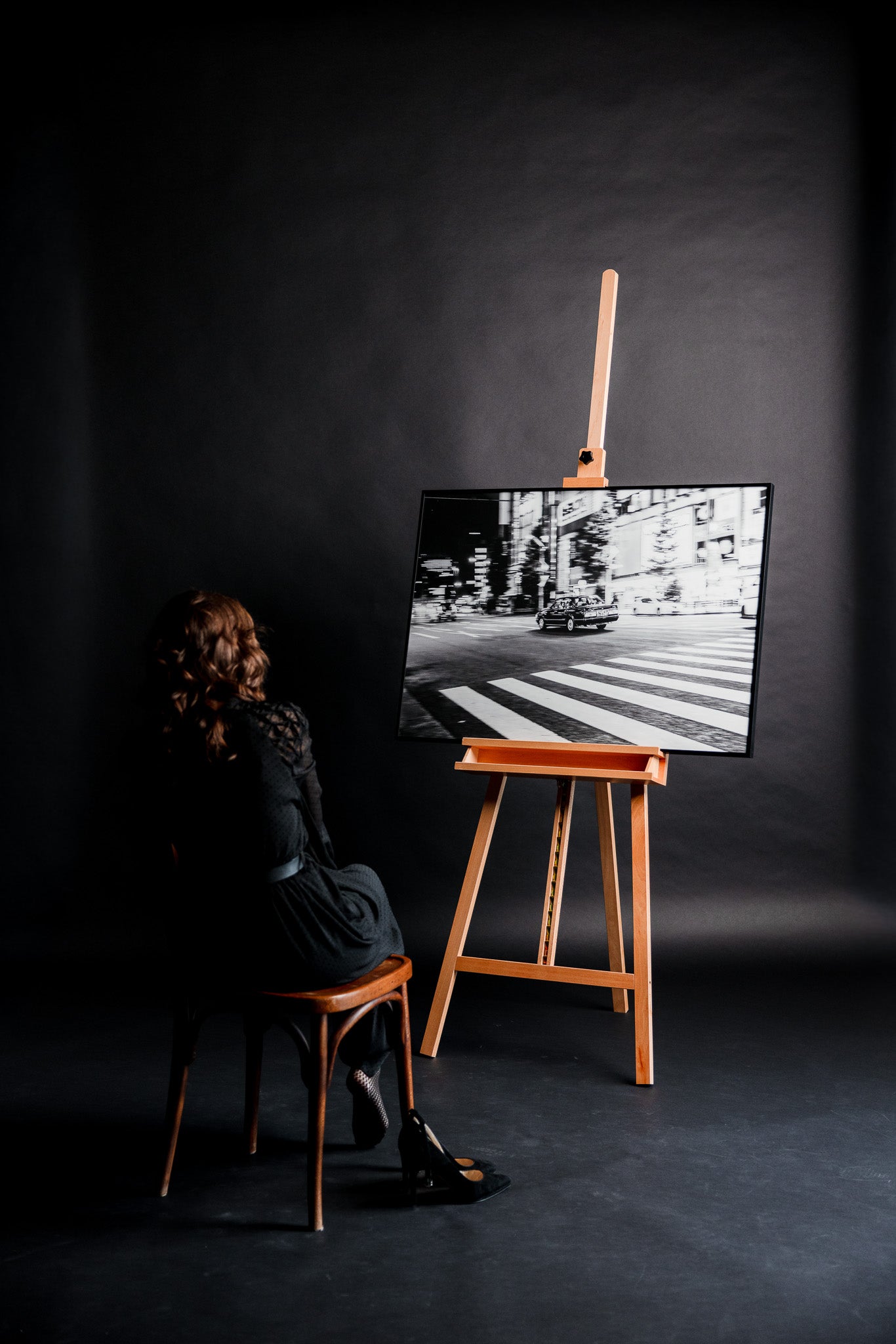 Schwarz-weiss Fotokunst „Just Got It“ von Motel June auf einer Staffelei, Panning Shot eines japanischen Taxis an einer Kreuzung in Tokyo mit weich verzerrten Zebrastreifen, elegante Frau sitzt auf einem alten Stuhl und betrachtet das Bild im Studio.
