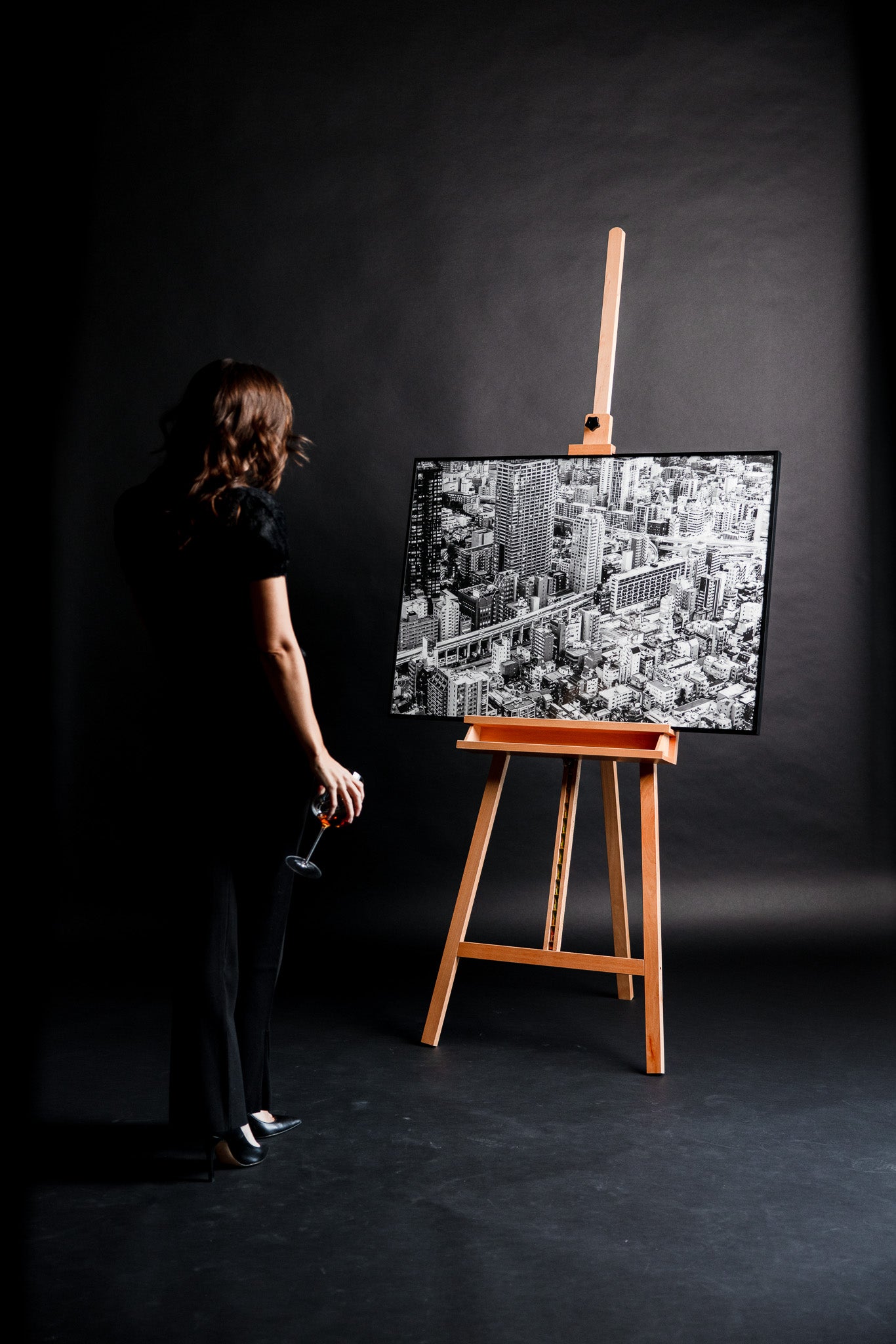 Schwarz-weiss Fotografie „Neighbourhood“ von Motel June auf einer Staffelei, Blick auf Downtown Tokyo von oben, junge Frau in schwarzem eleganten Outfit steht davor und betrachtet das Kunstwerk mit einem locker gehaltenen Glas Champagner im Fotostudio.