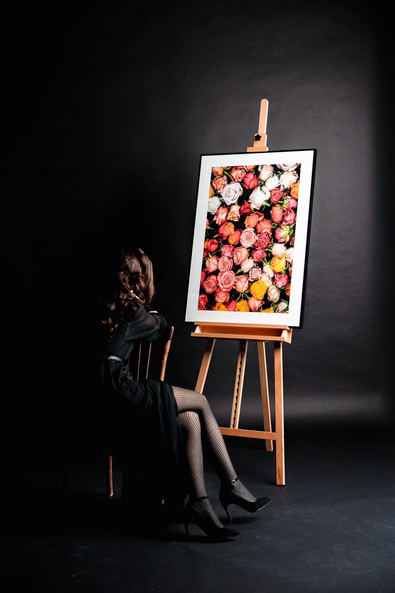 Farbige Fine Art Fotografie „Roses With Thorns“ von Motel June auf einer Staffelei, aufgenommen in Zürich mit einem Meer aus farbigen Rosen, junge Frau in schwarzer eleganter Kleidung sitzt mit verschränkten Beinen auf einem braunen Stuhl und betrachtet das Kunstwerk im Fotostudio.