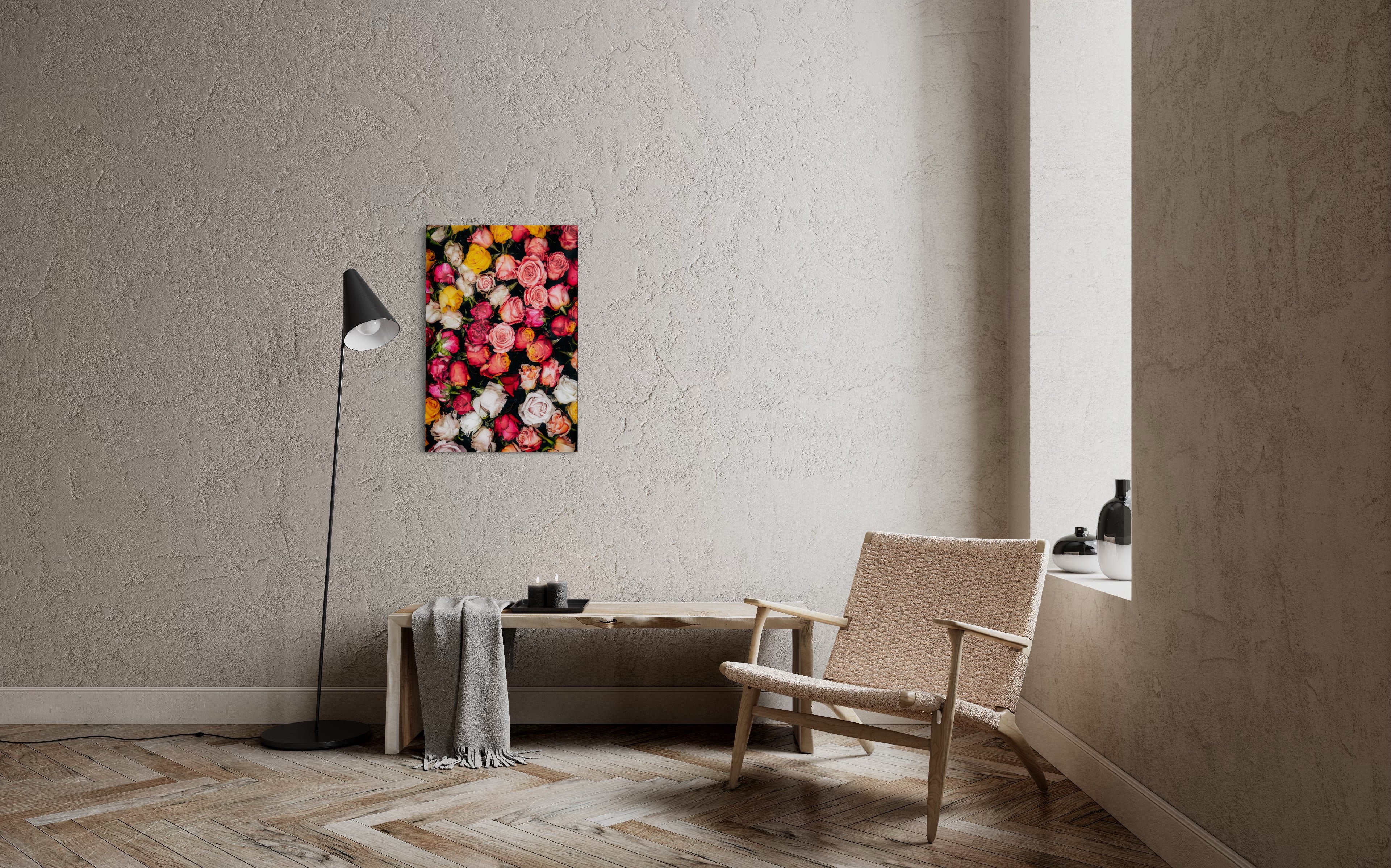 Der Fine Art Print "Roses with Thorns" hängt an einer weissen Betonwand; das farbenreiche Meer aus gelben, roten, pinken und weißen Rosen vor schwarzem Hintergrund wird links von einer Lampe und rechts von einem modernen Sessel eingerahmt.