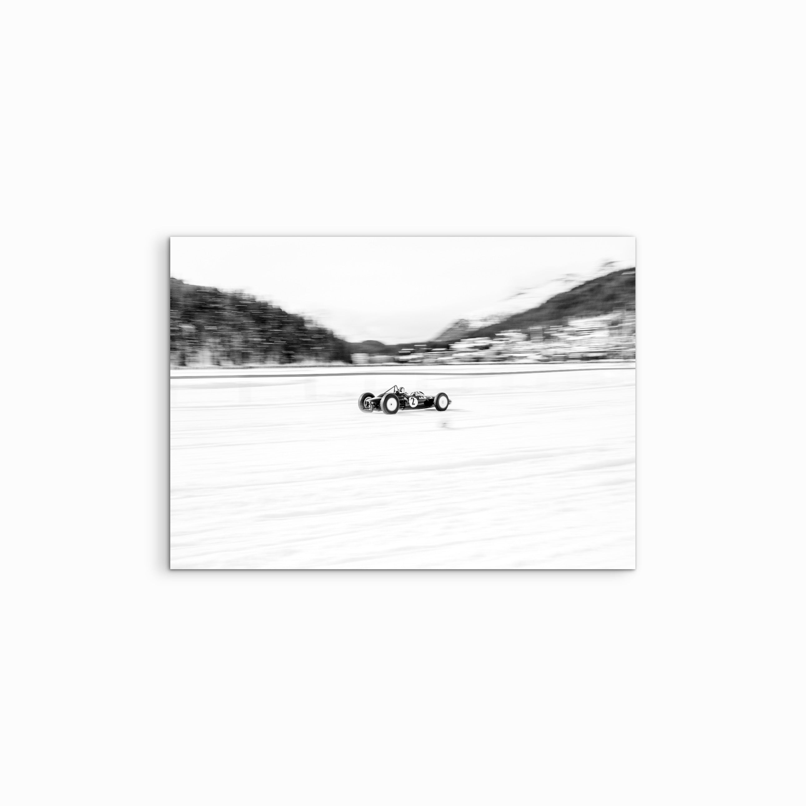Vor einem weissen Hintergrund zeigt der Fine Art Print „Flying Lotus“ eine schwarz-weisse Aufnahme eines vintage F1-Lotus, der über den vereisten und verschneiten See von St. Moritz rast, umgeben von winterlichen Bäumen und eingefangen in einer dynamischen Motorsport-Szene.