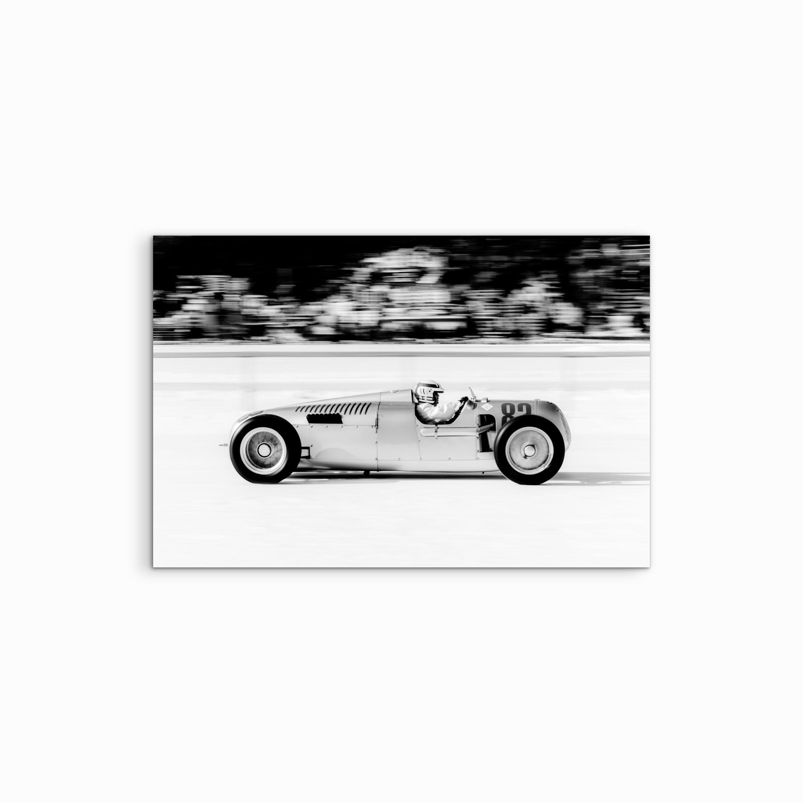Der Fine Art Print "A Flash" hängt vor einem weissen Hintergrund und zeigt eine Schwarz-Weiss-Fotografie eines vorbeirasenden Vintage-Rennwagens im Schnee mit dynamisch verzerrtem Hintergrund.