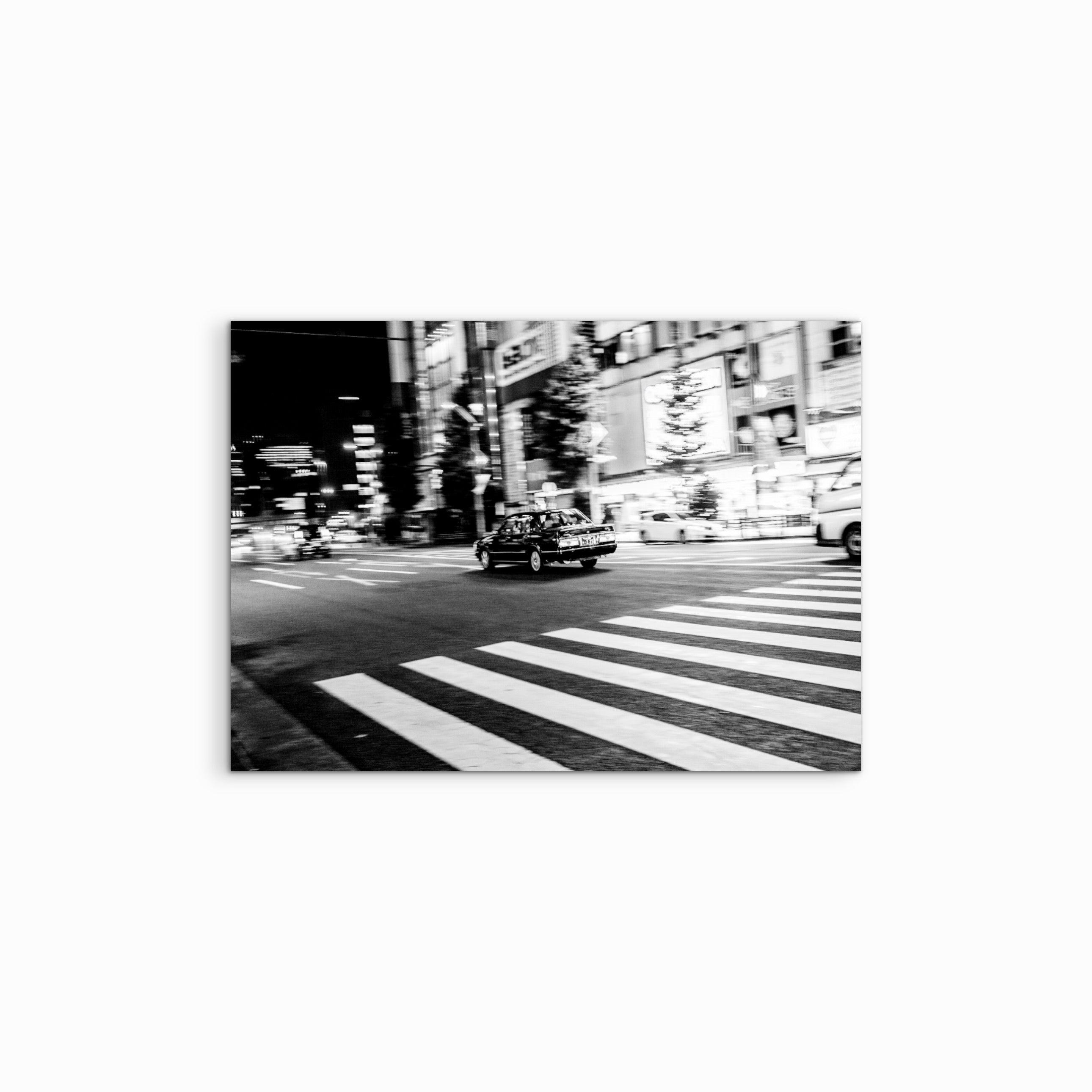 Vor einem weissen Hintergrund hängt der Fine Art Print „Just Got It“, der in einer kontrastreichen Schwarz-Weiss-Fotografie ein vorbeifahrendes Auto auf Tokios hell beleuchteter Strasse nahe eines Fussgängerstreifens zeigt und das dynamische Nachtleben der Stadt widerspiegelt.