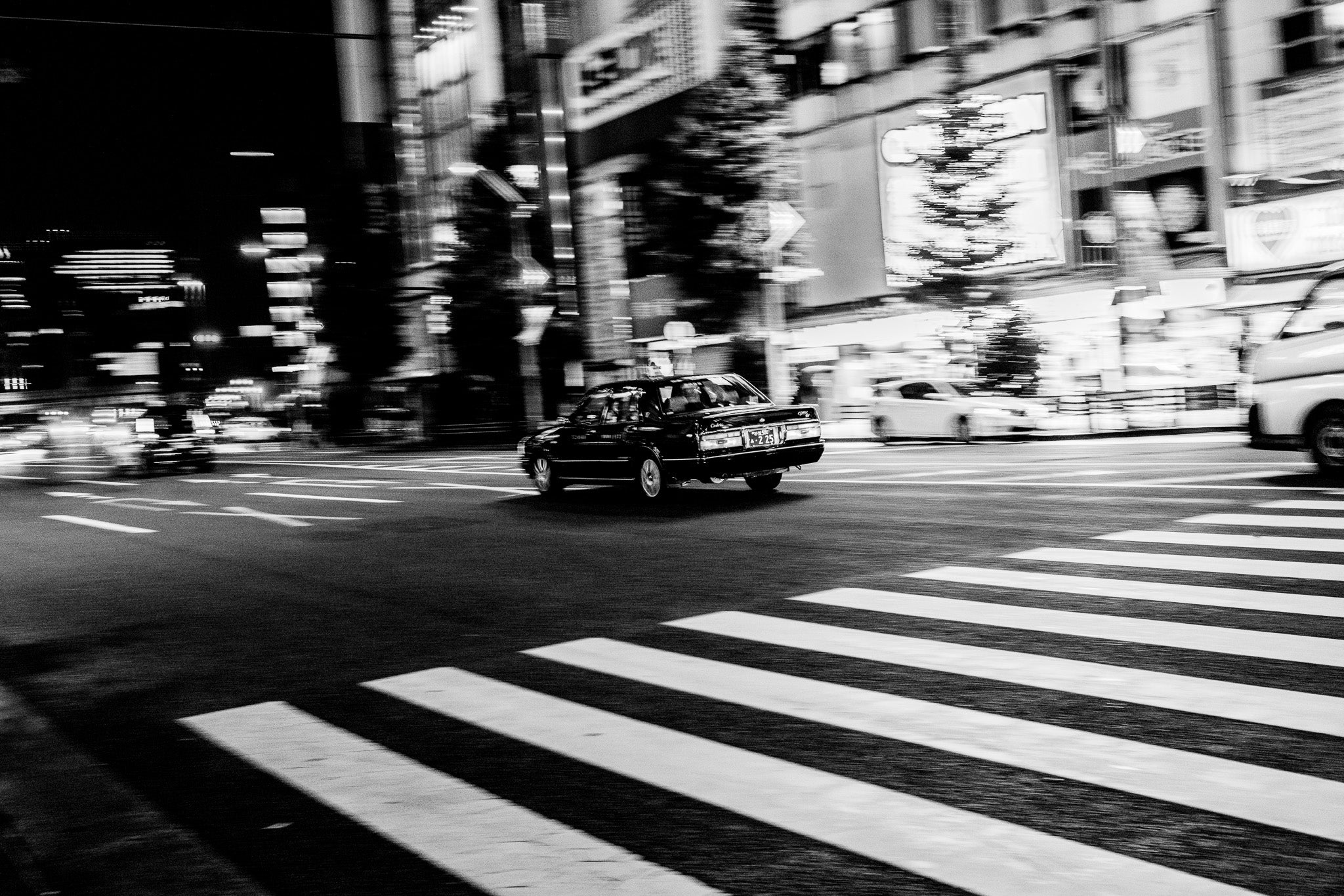 Schwarzes Auto auf einer hell beleuchteten Strasse neben einem weissen Fußgängerstreifen in Tokio, aus dem Schwarz-Weiss Fine Art Print "Just got it".