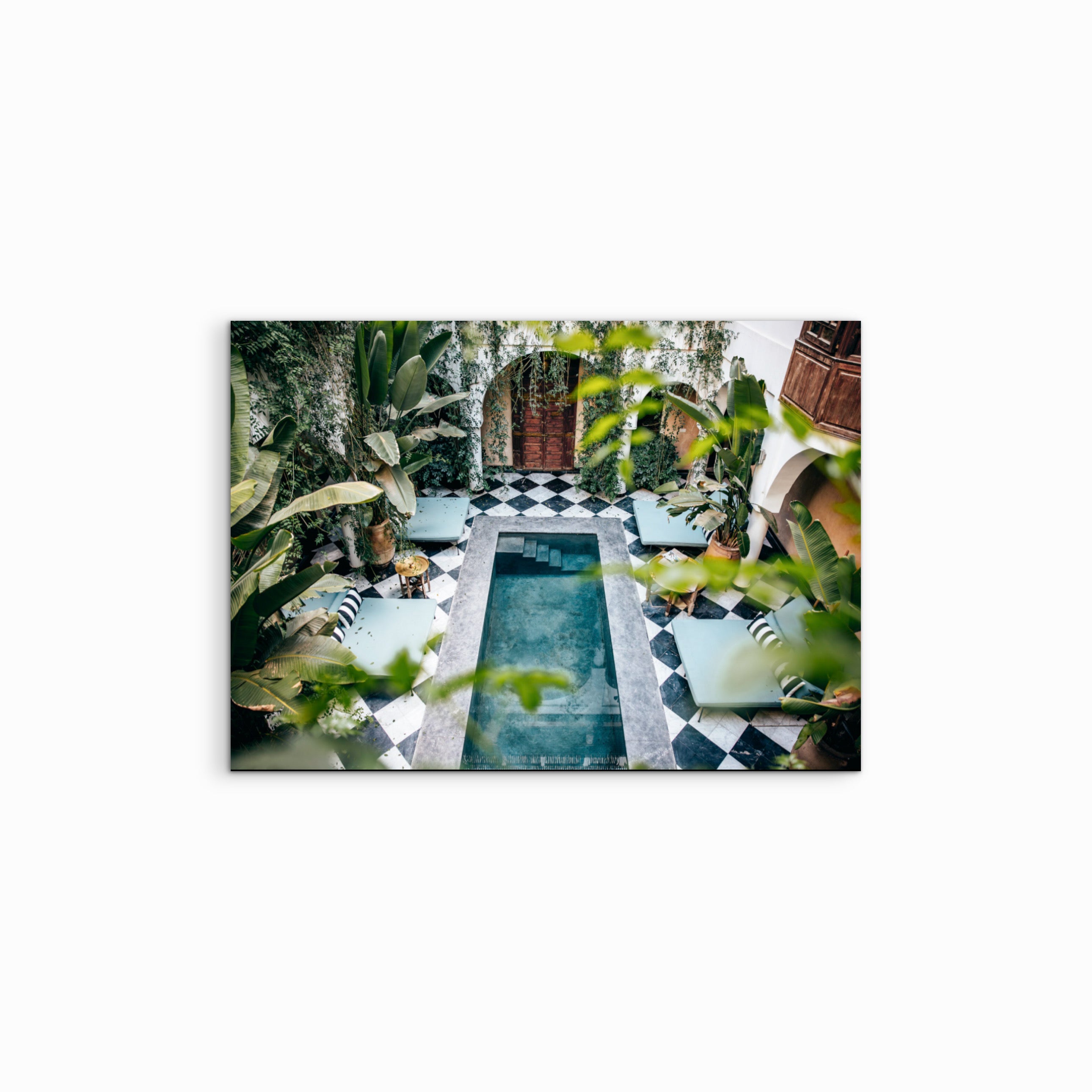 Der Fine Art Print „The Oasis“ zeigt das eindrucksvolle Innere eines Riads: Vom oberen Balkon blickt man auf einen grossen quadratischen Pool mit klarem blauem Wasser, umgeben von schwarz-weiss gemusterten Mosaikfliesen, vier grossen Poolliegen und üppigen grünen Pflanzen, die die Wände überwachsen, während am Ende eine massive Holztür in ein Zimmer führt.